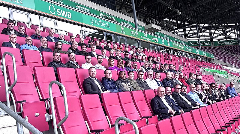 Eine große Gruppe von Männern sitzt in Reihen auf roten Stadionstühlen. Sie blicken in die Kamera, während im Hintergrund Werbebanner und die Stadionarchitektur zu sehen sind. Die Atmosphäre wirkt freundlich und einladend.