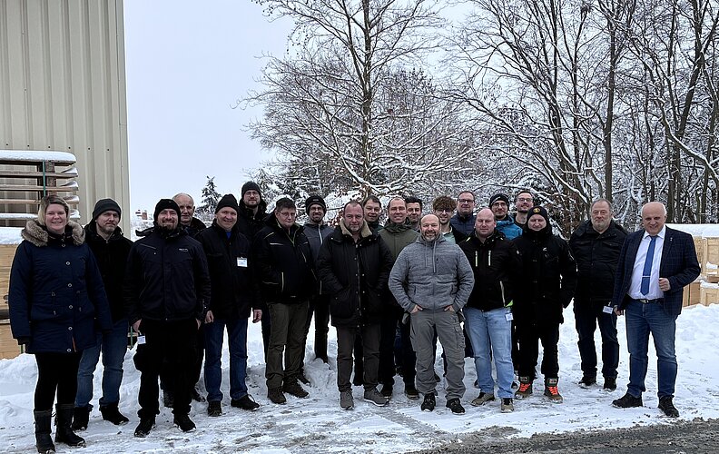 Eine Gruppe von etwa 25 Personen steht lächelnd im Schnee vor einem Gebäude. Einige tragen warme Winterkleidung. Bäume sind im Hintergrund sichtbar, und die Atmosphäre wirkt freundlich und gemeinschaftlich.