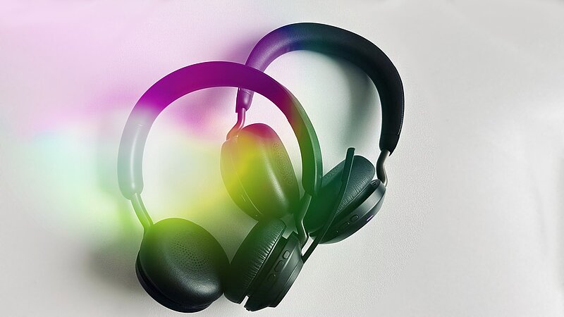 Zwei Headsets liegen auf einer hellen Oberfläche. Eines ist schwarz und das andere hat eine grüne Farbe. Im Hintergrund sind bunte Lichteffekte zu sehen, die dem Bild eine dynamische Note verleihen.