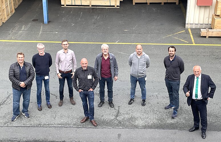 Eine Gruppe von neun Männern steht auf einem gepflasterten Platz vor einem Lagerhaus. Sie posieren in zwei Reihen, einige lächeln, während andere neutral schauen. Im Hintergrund sind Holzkisten sichtbar. Es ist ein klarer Tag.