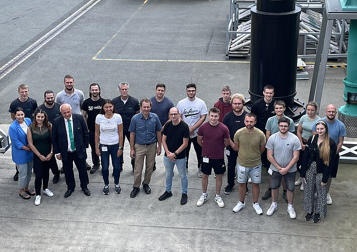 Eine Gruppe von etwa 25 Personen steht auf einem Gehweg in einem Außenbereich. Die Teilnehmer sind gemischt, einige tragen Arbeitskleidung, andere Freizeitkleidung. Im Hintergrund sind industrielle Anlagen und Ausrüstungen sichtbar. Die Gruppe lächelt und posiert gemeinsam für das Foto.