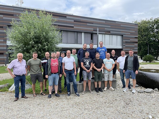 Gruppenfoto von 18 Männern im Freien, vor einem modernen Gebäude mit einer Grünfläche im Hintergrund. Einige tragen Shirts, Shorts und Sandalen, andere T-Shirts und Sneakers. Die Stimmung scheint freundlich und entspannt zu sein.