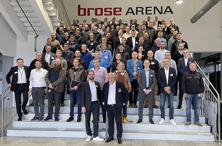 Gruppennfoto von über 100 Personen auf einer Treppe in der Brose Arena. Die Teilnehmer sind überwiegend Männer, gekleidet in formeller oder Freizeitkleidung. Einige tragen Namensschilder. Die Atmosphäre ist freundlich und kollegial.