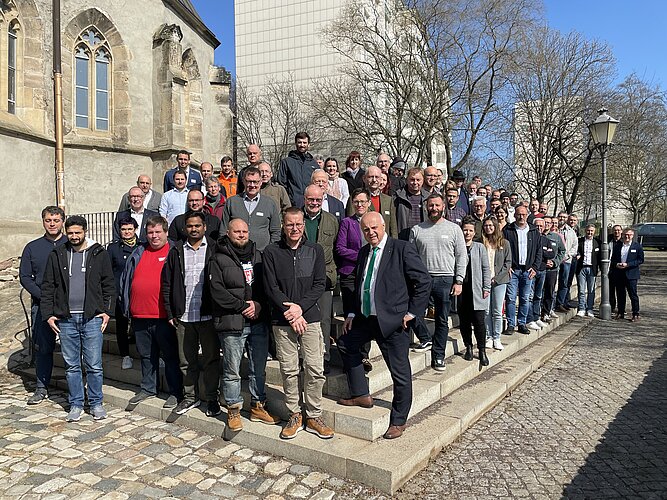 Gruppensfoto von etwa 60 Personen, die auf Treppen an einer historischen Gebäudegruppe stehen, umgeben von Bäumen und modernen Gebäuden. Die Personen lächeln und posieren für das Foto, viele tragen legere Kleidung.