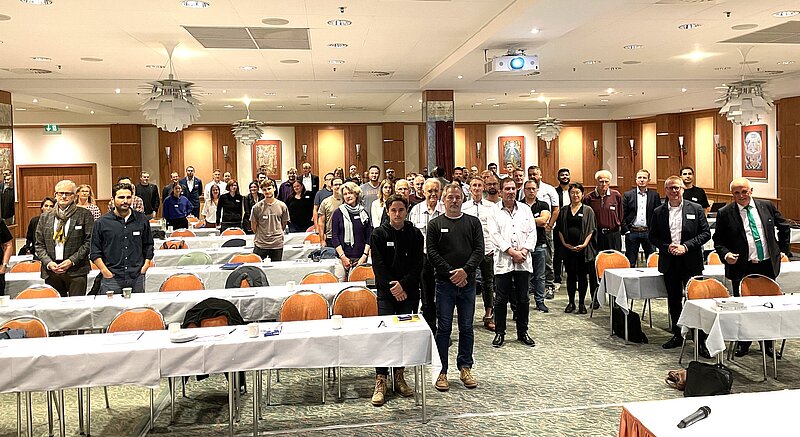 Eine Gruppe von Menschen steht in einem Konferenzraum. Tische mit weißen Tischdecken sind im Hintergrund angeordnet. Die Teilnehmer sind angezogen, einige haben Namensschilder. Der Raum ist beleuchtet und es sind Projektoren und andere Konferenzelemente sichtbar.