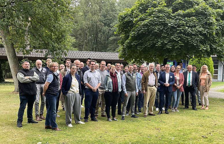 Eine Gruppe von etwa 30 Personen steht auf einer grünen Wiese, umgeben von Bäumen und einem Gebäude im Hintergrund. Die Menschen lächeln und posieren für ein Gruppenfoto, gekleidet in verschiedene Freizeit- und Geschäftsoutfits.