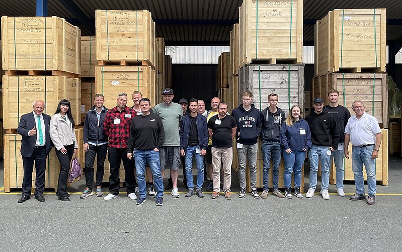 Eine Gruppe von 16 Personen posiert in einer Lagerhalle zwischen großen Holzkisten. Im Hintergrund sind viele Kisten gestapelt. Die Gruppe besteht aus Männern und Frauen, einige tragen Namensschilder. Es herrscht eine freundliche Atmosphäre.