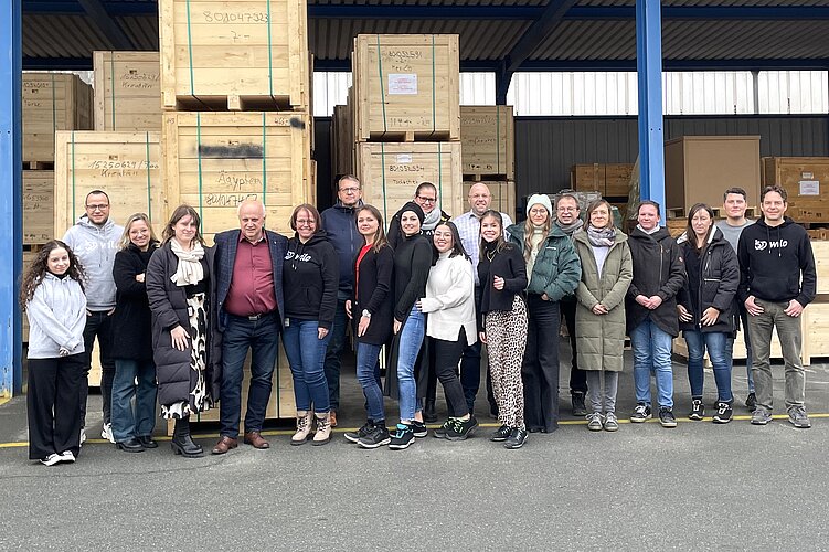 Eine Gruppe von 22 Personen steht vor großen Holzkisten in einem Lager. Sie lächeln und posieren gemeinsam für das Foto. Im Hintergrund sind weitere gestapelte Kisten sichtbar, und es herrscht ein klarer Himmel.