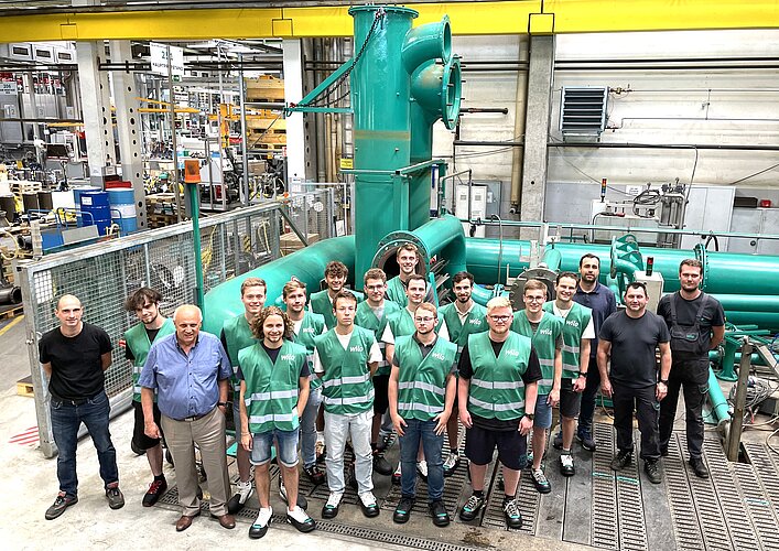 Eine Gruppe von jungen Menschen in grünen Arbeitswesten steht vor großen Maschinen in einer Fabrik. Im Hintergrund sind weitere Maschinen und Ausrüstungen sichtbar. Die Personen lächeln und posieren für das Foto.