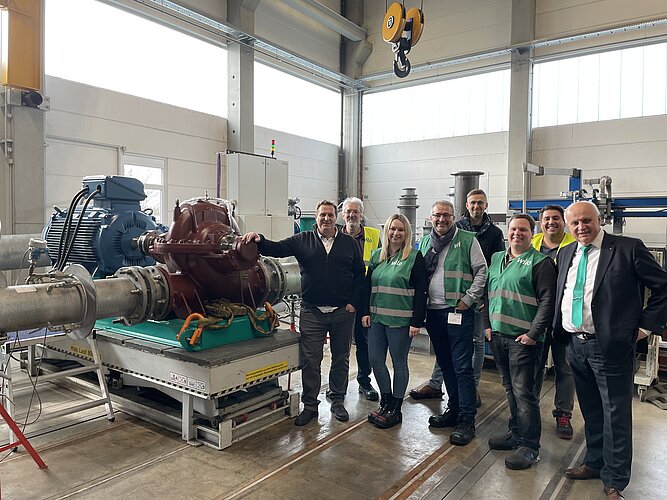 Eine Gruppe von acht Personen steht in einer Industriehalle vor einer großen Maschine. Einige tragen grüne Sicherheitswesten. Im Hintergrund sind Rohre und eine weitere Maschine zu sehen. Die Atmosphäre wirkt freundlich und professionell.