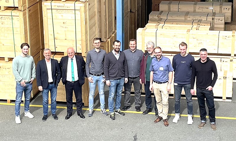 Eine Gruppe von zehn Männern steht in einer Lagerhalle vor großen Holzverpackungen. Sie lächeln und posieren für das Foto. Die Männer tragen verschiedene, legere Kleidung. Im Hintergrund sind ordentlich gestapelte Holzpaletten sichtbar.