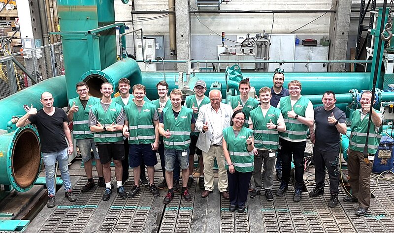 Eine Gruppe von zwölf Personen steht in einer industriellen Umgebung, umgeben von großen grünen Rohren. Alle tragen auffällige Warnwesten. Sie zeigen mit beiden Händen Daumen hoch und lächeln in die Kamera. Im Hintergrund sind Maschinen und Arbeitsmaterialien sichtbar.
