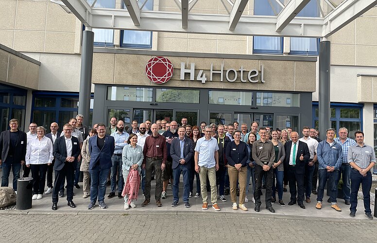 Eine große Gruppe von Menschen steht vor dem H4 Hotel. Sie lächeln und posieren für das Foto. Die Gruppe besteht aus Männern und Frauen unterschiedlichen Alters, die in formeller und sportlicher Kleidung gekleidet sind.
