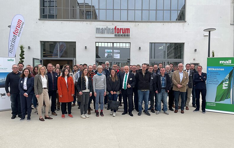 Eine große Gruppe von Menschen steht vor dem Eingang zum marlinforum. Sie lächeln und posieren für ein Gruppenfoto. Im Hintergrund sind Banner und Beschilderungen sichtbar, die mit den Worten "Willkommen" und Firmenlogos beschriftet sind.