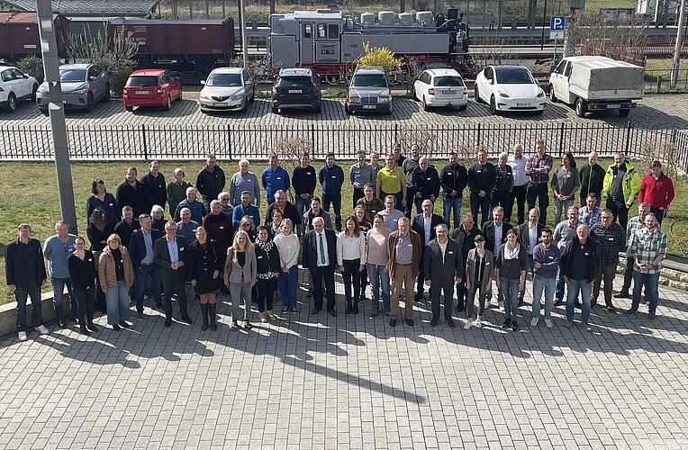 Eine große Gruppe von Menschen steht auf einem gepflasterten Platz, umgeben von Autos und einer Eisenbahn im Hintergrund. Die Personen wirken freundlich und sind unterschiedlich gekleidet. Es ist ein sonniger Tag.