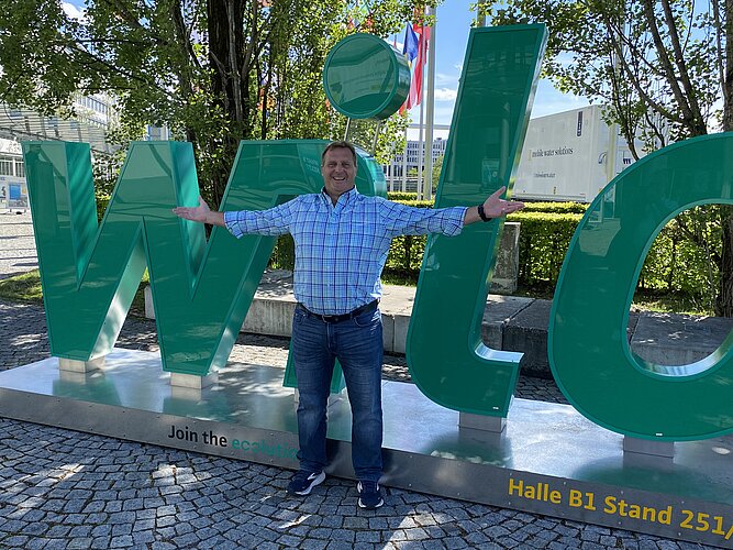 Ein lächelnder Mann steht vor großen, grünen Buchstaben, die das Wort "wild" bilden. Er hat die Arme ausgebreitet und trägt ein kariertes Hemd. Im Hintergrund sind Bäume und Flaggen sichtbar.