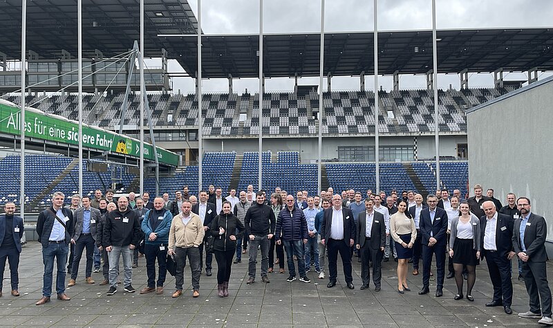 Eine große Gruppe von Menschen steht auf einem Platz vor einem Stadion. Die Anwesenden lächeln und posieren für das Foto. Im Hintergrund sind leere Sitzreihen des Stadions zu sehen, während ein Teil des Daches darüber sichtbar ist.