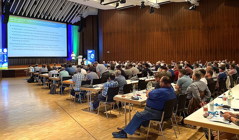 Eine große Gruppe von Teilnehmern sitzt in einem Konferenzraum mit Sitzplätzen an Tischen. Auf einer Leinwand wird eine Präsentation gehalten, während ein Redner auf der Bühne spricht. Die Atmosphäre ist professionell und konzentriert.