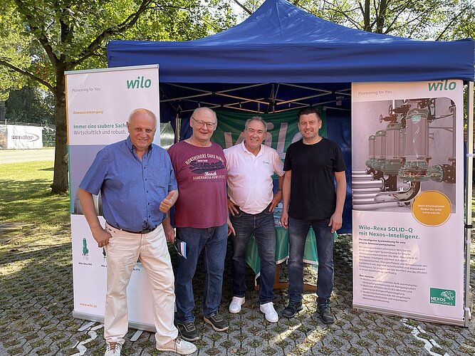 Vier Männer stehen vor einem blauen Zelt mit Werbeflächen für die Firma Wilo. Sie lächeln und posieren für das Foto, umgeben von Bäumen und einem hellen, sonnigen Tag.