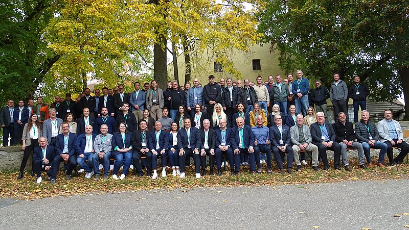 Eine große Gruppe von Menschen steht und sitzt vor Bäumen in einer farbenfrohen herbstlichen Umgebung. Die Personen sind in verschiedenen Kleidungsstücken gekleidet und tragen Namensschilder. Einige schauen in die Kamera, während andere sich unterhalten.