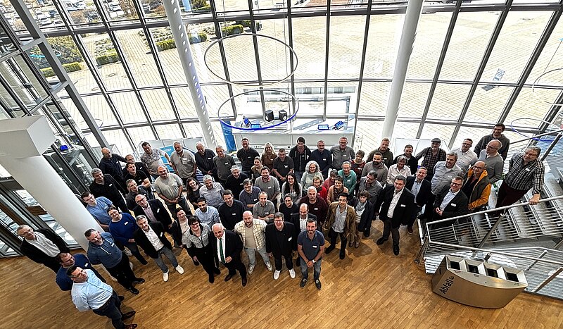 Eine große Gruppe von Menschen steht in einem modernen, lichtdurchfluteten Raum mit Glaswänden. Sie posieren gemeinsam und schauen in die Kamera. Der Boden ist aus Holz, und im Hintergrund sind Fenster und eine Treppe sichtbar.