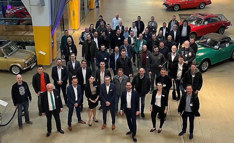 Eine große Gruppe von über 50 Personen steht in einem Auto-Museum. Im Hintergrund sind mehrere klassische Fahrzeuge zu sehen. Die Anwesenden tragen verschiedene Business-Outfits und lächeln in die Kamera. Die Atmosphäre wirkt freundlich und einladend.