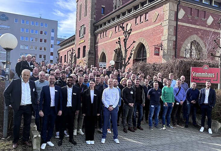 Eine große Gruppe von Menschen steht vor einem historischen Gebäude. Sie tragen verschiedene Kleidung und lächeln in die Kamera. Im Hintergrund sind Bäume und ein Schild mit der Aufschrift "Stammhaus" sichtbar. Die Szene vermittelt ein Gefühl von Gemeinschaft und Zusammengehörigkeit.