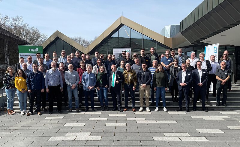 Eine Gruppe von etwa 60 Personen, darunter Männer und Frauen, steht auf einer Treppe vor einem modernen Gebäude mit einem auffälligen Dachdesign. Alle Teilnehmer lächeln und posieren für das Foto an einem sonnigen Tag.