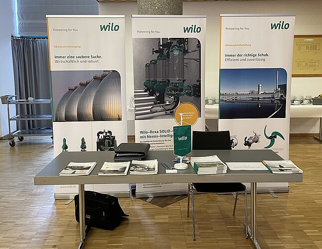 Ein Messestand mit drei Banner von Wilo, die Pumpen und technische Lösungen präsentieren. Auf dem Tisch liegen Informationsmaterialien und Broschüren, sowie ein Tischaufsteller mit dem Wilo-Logo. Im Hintergrund sind Tische und eine Wand zu sehen.