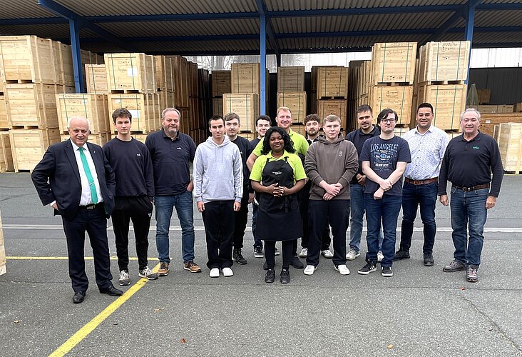 Eine Gruppe von 15 Personen steht in einer Lagerhalle mit Holzpaletten im Hintergrund. Sie lächeln und sind in Arbeitskleidung oder Freizeitkleidung gekleidet. Einige Personen tragen Schürzen, während sie zusammen posieren.