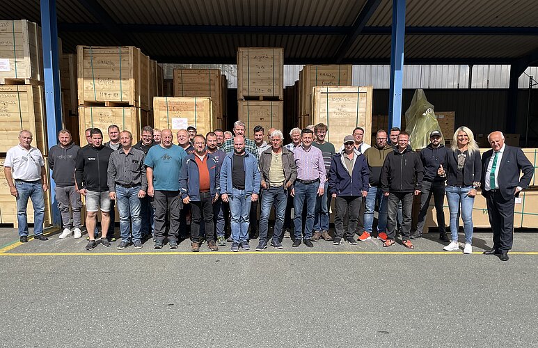 Eine Gruppe von etwa 30 Personen steht vor einem Lagerhaus mit Holzpaletten. Die Teilnehmenden sind Männer und Frauen unterschiedlichen Alters, lächelnd und in legerer Kleidung. Im Hintergrund sind große, gestapelte Kisten sichtbar.