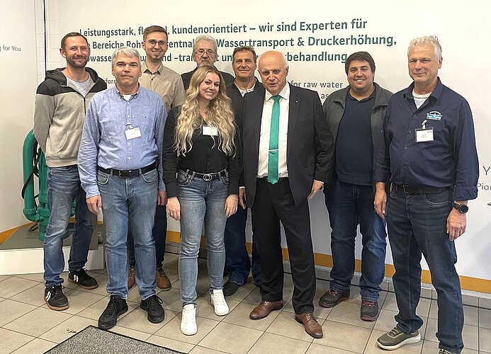 Eine Gruppe von zehn Personen posiert auf einem Event. Sie stehen vor einer Wand mit Text und Logos. Die Männer tragen verschiedene Kleidung, von lässig bis formell, während eine Frau in der Mitte lächelt. Ein Mann trägt einen grünen Krawatte.