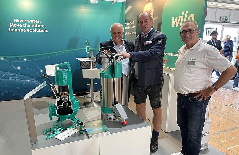 Drei Männer stehen an einem Messestand von Wilo, umgeben von Maschinen zur Wasserförderung. Im Vordergrund sind zwei Pumpenmodelle und Zubehörteile zu sehen. Der Stand hat ein modernes Design mit einer großen Wilo-Logo und einem futuristischen Hintergrund.