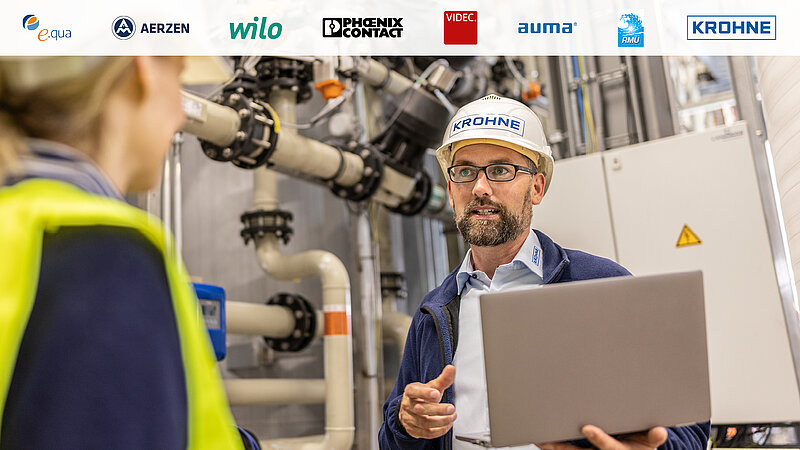 Ein Mann in einem weißen Helm und Brille spricht mit einer Frau, die eine Warnweste trägt. Er hält einen Laptop in der Hand. Im Hintergrund sind Rohrleitungen und Maschinen sichtbar. Oben sind Unternehmenslogos zu sehen.