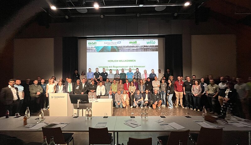 Eine Gruppe von Personen steht auf einer Bühne vor einem Bildschirm mit der Aufschrift "HERZLICH WILLKOMMEN" und dem Thema der Veranstaltung. Im Vordergrund sind Tische mit Wasserflaschen und Informationsmaterial. Die Teilnehmer sind in verschiedenen Outfits und stehen in mehreren Reihen.