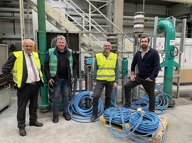 Vier Männer stehen in einer Industrieumgebung, alle tragen Sicherheitswesten. Im Hintergrund sind Rohre und technische Ausrüstung zu sehen, darunter grüne Rohre und Kabel. Auf dem Boden liegen mehrere blaue Kabel.