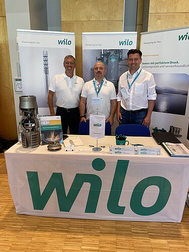 Drei Männer stehen vor einem Messestand mit einem großen „wilo“-Logo. Auf dem Tisch sind verschiedene Produkte, Informationsmaterialien und ein Pumpenmodell ausgelegt. Im Hintergrund sind Werbetafeln mit technischen Informationen und Bildern zu sehen.