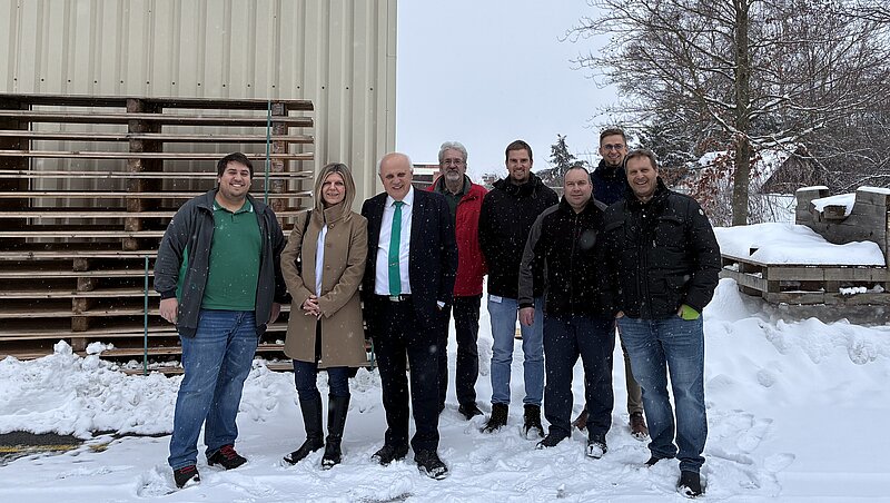 Eine Gruppe von neun Personen steht im Schnee vor einem Gebäude. Einige tragen Winterkleidung, und eine Person hält eine grüne Krawatte. Im Hintergrund sind Holzpaletten und schneebedeckter Boden sichtbar.