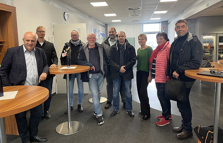 Eine Gruppe von neun Personen steht in einem hellen Raum mit Tischen. Sie sind unterschiedlich gekleidet und lächeln in die Kamera. Im Hintergrund sind Gegenstände und Automaten sichtbar, die auf eine Büroumgebung hinweisen. Es gibt einen großen Fensterbereich, der Tageslicht hereinlässt.
