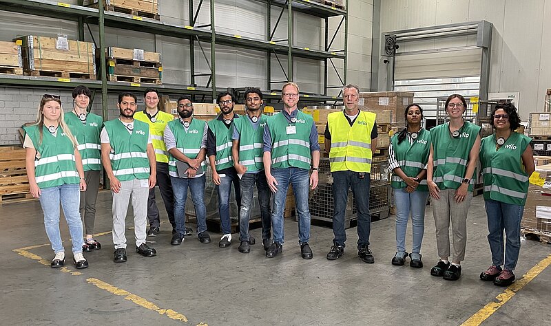 Gruppenfoto von 13 Personen in einem Lager. Die meisten tragen grüne Westen, einige auch gelbe Warnwesten. Sie stehen zusammen auf einem gepflasterten Boden vor Regalen mit Kartons im Hintergrund. Alle blicken zur Kamera und lächeln.