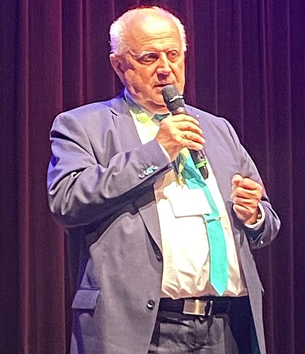 Ein älterer Mann mit grauen Haaren steht auf einer Bühne und spricht ins Mikrofon. Er trägt einen blauen Anzug und ein weißes Hemd mit einer farbigen Krawatte. Im Hintergrund sind Vorhänge zu sehen, die die Bühne beleuchten.