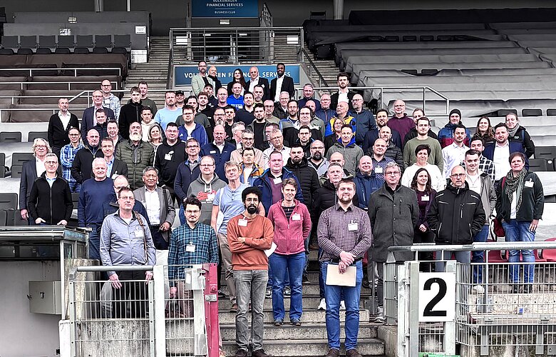 Eine große Gruppe von Menschen steht in einer Sportarena. Die Personen sind unterschiedlich gekleidet und stehen in mehreren Reihen zusammen. Einige halten Papiere oder Ordner. Im Hintergrund sind leere Sitzplätze zu sehen.