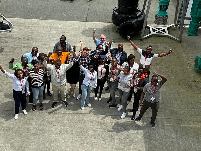 Eine Gruppe von etwa 20 Menschen steht im Freien und hebt die Arme in die Luft. Alle scheinen glücklich und feiern einen gemeinsamen Moment. Die Kleidung variiert, einige tragen T-Shirts, andere Hemden und Blusen. Im Hintergrund sind industrielle Anlagen sichtbar.