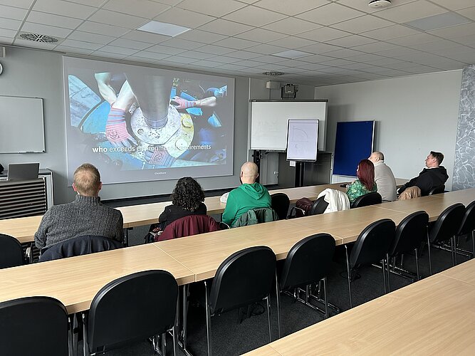 Eine Gruppe von Personen sitzt in einem Konferenzraum und schaut auf eine Präsentation, die auf eine Leinwand projiziert wird. Das Thema scheint sich um technische oder mechanische Elemente zu drehen. Die Atmosphäre ist konzentriert und die Teilnehmer hören aufmerksam zu.
