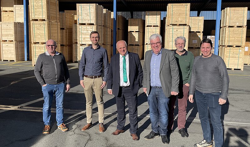 Eine Gruppe von sechs Männern steht vor großen Holzpaletten in einem Lager. Sie lächeln in die Kamera und tragen formelle sowie legere Kleidung. Die Umgebung sieht industriell aus.