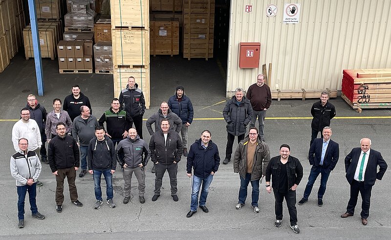 Eine Gruppe von 20 Männern steht in einem Außenbereich vor einem Lager mit Holzregalen. Einige Personen tragen Arbeitskleidung, andere Freizeitkleidung. Im Hintergrund sind große Holzkisten und ein Lagergebäude zu sehen. Die Stimmung wirkt freundlich und kollegial.