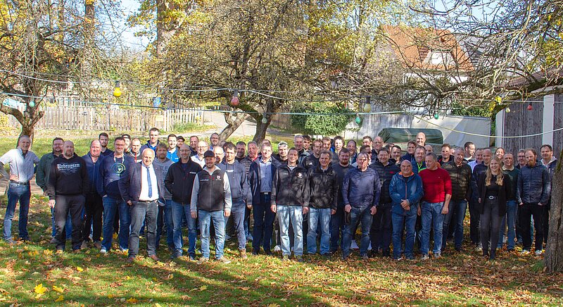 Eine große Gruppe von Männern und Frauen steht im Freien auf einer Wiese, umgeben von Bäumen mit herbstlichen Blättern. Sie lächeln und posieren gemeinsam für ein Foto, während die Sonne scheint.