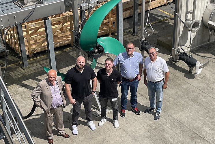 Fünf Personen stehen neben einem großen grünen Propeller in einer industriellen Umgebung. Sie tragen unterschiedliche Kleidung und lächeln in die Kamera. Im Hintergrund sind weitere technische Geräte und Materialien zu sehen.