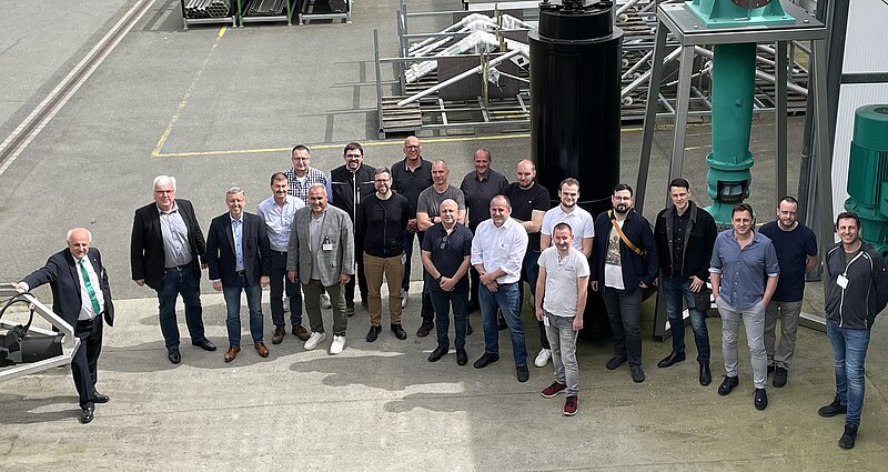 Eine Gruppe von etwa 20 Menschen steht im Freien auf einem Industriegelände. Sie posieren gemeinsam vor großen Maschinen und Ausrüstungen. Die Personen sind lässig gekleidet und lächeln. Im Hintergrund sind metallische Strukturen und Ausrüstungen sichtbar.