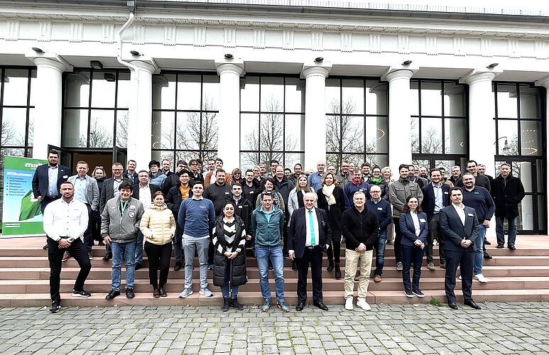 Eine große Gruppe von Menschen steht auf den Stufen eines Gebäudes. Die Teilnehmer lächeln und posieren für ein Foto. Im Hintergrund sieht man große Fenster und Bäume. Die Szene vermittelt einen festlichen Anlass oder ein gemeinsames Treffen.